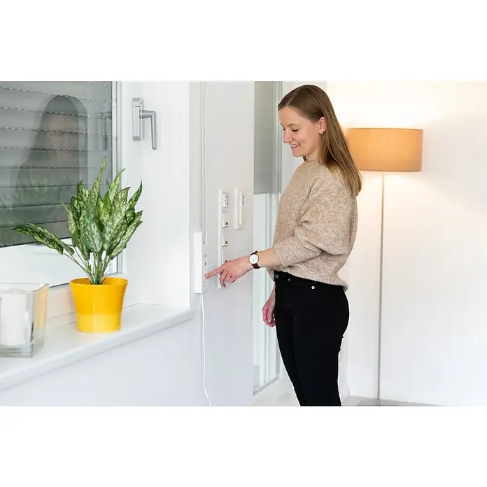 Schellenberg Eje octogonal telescópico - Apto para: Mini sistemas de persianas enrollables - Una mujer acciona un control de persiana en una pared junto a una ventana con una planta en maceta y una lámpara de pie al fondo.