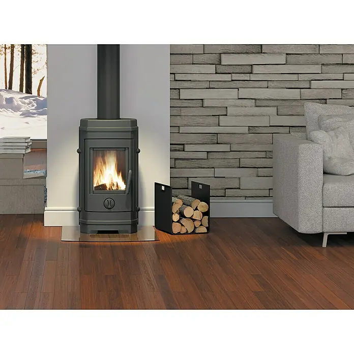 Invicta Estufa de leña Remilly - 7 kW, Volumen de calefacción: 210 m³, Antracita - Una estufa de leña con fuego crepitante se asienta sobre una base de vidrio, junto a un leñero, creando un ambiente cálido y acogedor.