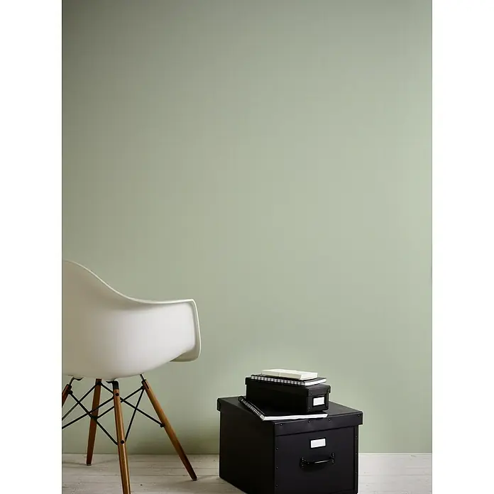 A.S. Création Greenery Vliestapete - Hellgrün, Uni, 10,05 x 0,53 m - Wand mit mintfarbener Farbe, neben einem weißen Stuhl mit Holzbeinen und schwarzen Schachteln mit Büchern darauf.