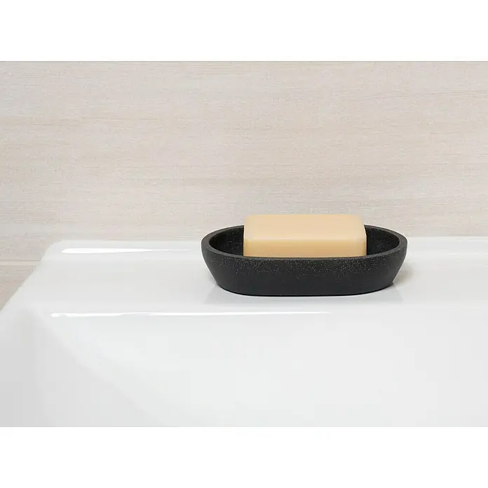 Camargue Seifenschale Stone - Polyresin, Anthrazit, Länge: 13,1 cm - Nahaufnahme einer schwarzen Seifenschale mit einem Stück beiger Seife darauf, platziert auf einem weißen Waschbecken vor einer hellen Wand.