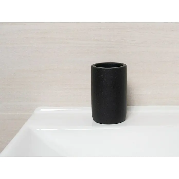 Camargue Zahnputzbecher Stone - Polyresin, Anthrazit, Höhe: 11,5 cm - Schwarzer, zylindrischer Zahnputzbecher auf einem weißen Waschtisch vor einer hellen, horizontal gestreiften Wand.