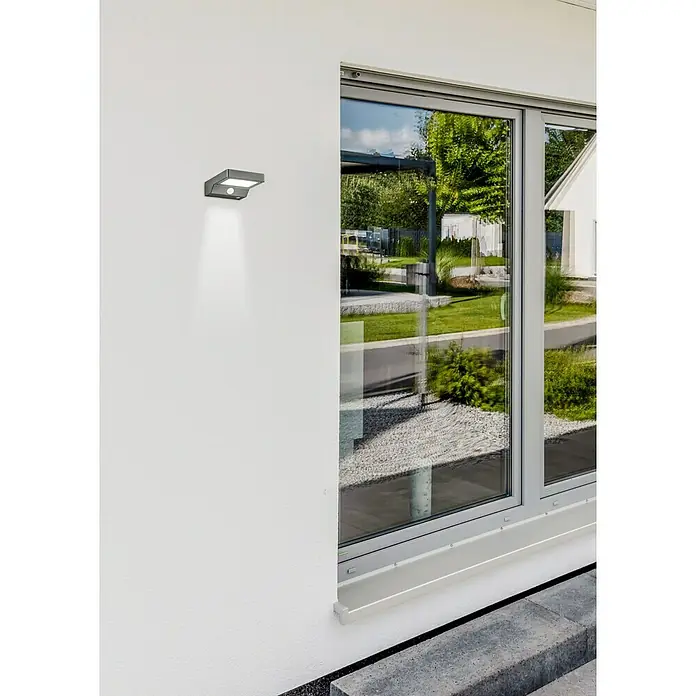 Reality Leuchten Solar-Außenwandleuchte Fomosa - 5 W, L x B x H: 16,5 x 12,5 x 4,5 cm, IP44, 340 lm - Eine moderne Außenleuchte an einer weißen Wand neben einem Fenster mit Blick auf einen Garten.