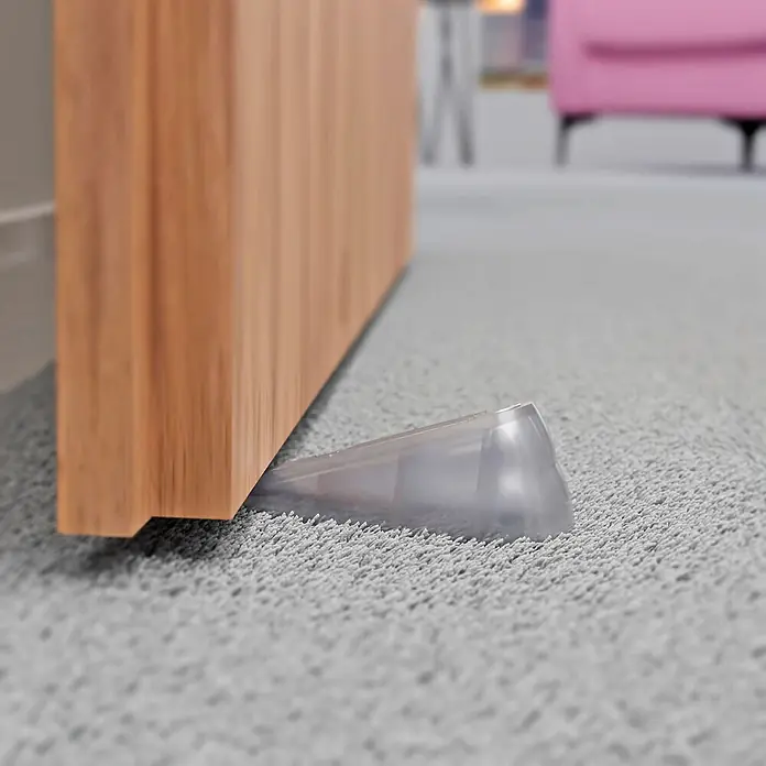 Walteco Türkeil - L x H: 100 x 36 mm, Material: Sonstiges - Ein transparenter Türstopper hält eine Holztür auf einem grauen Teppich offen und sorgt für sicheren Halt.