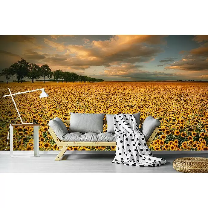 Papermoon Premium collection Fototapete Sonnenblumen - B x H: 450 x 280 cm, Vlies - Holzsofa mit grauen Kissen und gepunkteter Decke, neben moderner Lampe und geflochtenem Pouf, vor einer Sonnenblumenfeld-Wandmalerei.