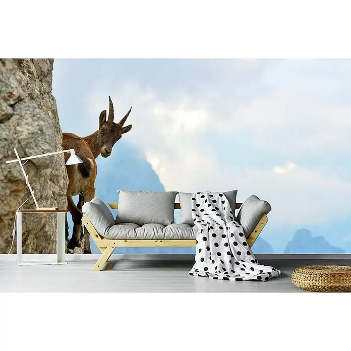 Papermoon Premium collection Fototapete Bergziege - B x H: 250 x 186 cm, Vlies - Ein helles Holzsofa mit grauen Kissen und einer gepunkteten Decke vor einem Wandbild mit einer Bergziege an einem Felsen und Himmelshintergrund.