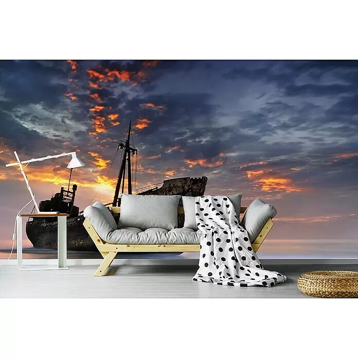Papermoon Premium collection Fototapete Der dunkle Reisende II - B x H: 200 x 149 cm, Vlies - Ein Holzsofa mit grauen Kissen und gepunkteter Decke vor einer Schiffswrack-Tapete.