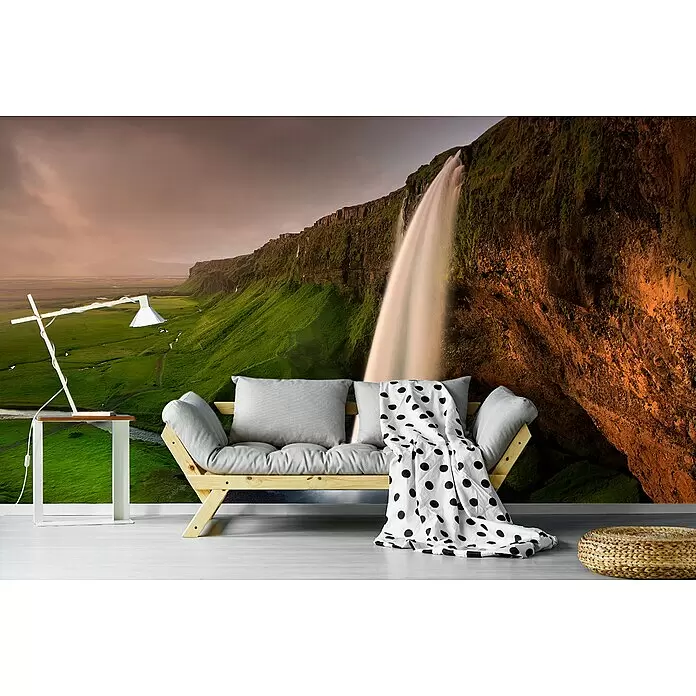 Papermoon Premium collection Fototapete Seljalandsfoss - B x H: 350 x 260 cm, Vlies - Ein helles Holzsofa mit grauen Kissen und gepunkteter Decke, ein weißer Beistelltisch mit Lampe und ein geflochtener Pouf im Wohnzimmer.