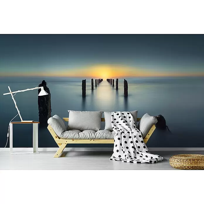 Papermoon Premium collection Fototapete Letztes Licht - B x H: 300 x 223 cm, Vlies - Helles Holzfutonsofa mit grauen Kissen und einer weiß-schwarz gepunkteten Decke, in einem Raum mit Meereswandbild. Eine Lampe und ein Beistelltisch sind in der Nähe.