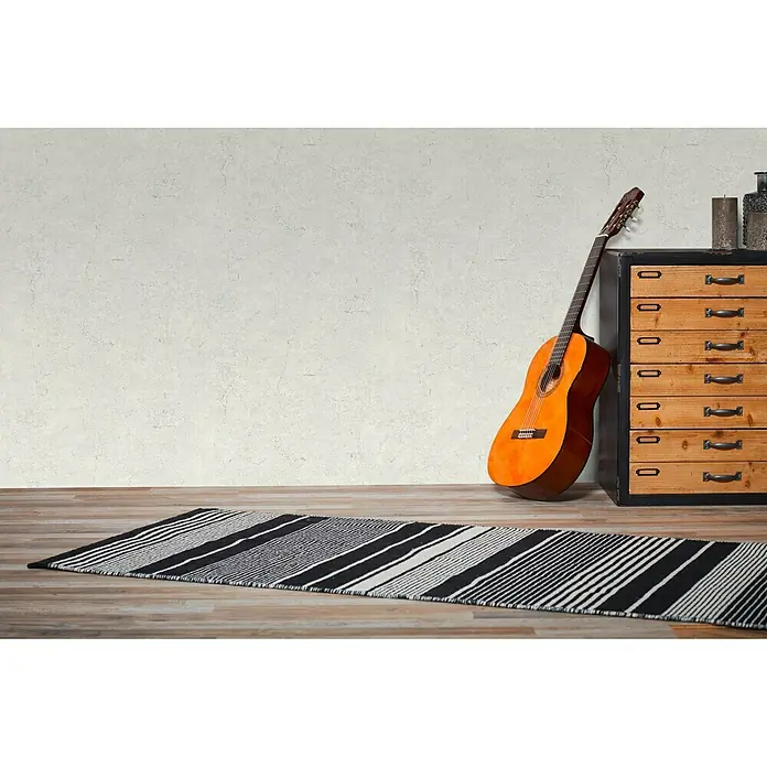A.S. Création Metropolitan Stories Vliestapete Betonoptik - Hellgrau, Betonoptik, 10,05 x 0,53 m - Ein gestreifter Teppich in Schwarz und Weiß liegt auf einem Holzboden, mit einer Gitarre und Kommode im Hintergrund.