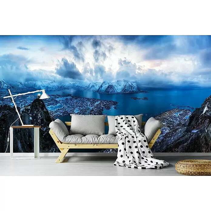 Papermoon Premium collection Fototapete Im Himmel auf Hazven - B x H: 200 x 149 cm, Vlies - Ein helles Holz-Futonsofa mit grauen Kissen und einer gepunkteten Decke, neben einem weißen Beistelltisch mit Lampe, vor einer Bergwandmalerei.