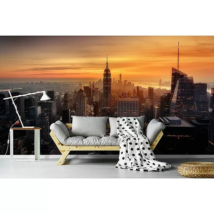Papermoon Premium collection Fototapete Manhattans Licht - B x H: 400 x 260 cm, Vlies - Helles Holz-Futonsofa mit hellgrauen Kissen und gepunkteter Decke in einem Raum mit New Yorker Skyline-Tapete bei Sonnenuntergang.