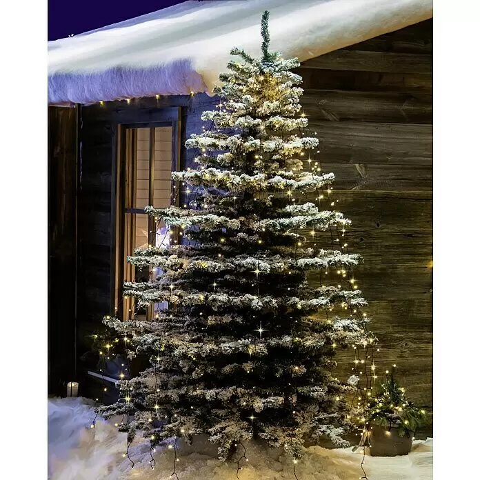 Konstsmide LED-Lichterkette Baummantel - Außen, Lichtfarbe: Bernstein, Anzahl Leuchtmittel: 400 Stk., Netzbetrieben - Ein künstlicher Weihnachtsbaum mit Kunstschnee und warmen Lichtern, draußen neben einer rustikalen Holzhütte.