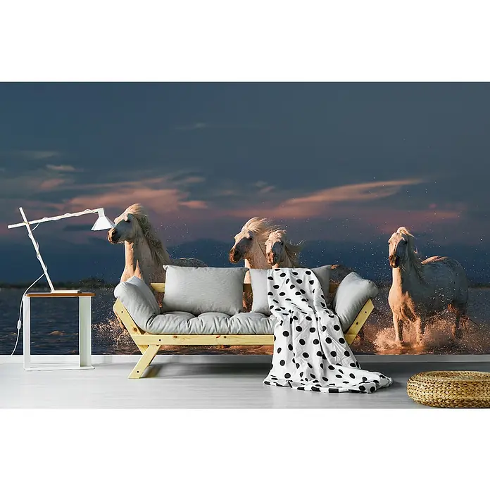 Papermoon Premium collection Fototapete Angels von Camargue im Sonnenrot - B x H: 400 x 260 cm, Vlies - Eine moderne Wohnraumgestaltung mit einer Fototapete, die eine Herde weißer Pferde im Wasser zeigt, ergänzt durch Sofa, Lampe und Hocker.