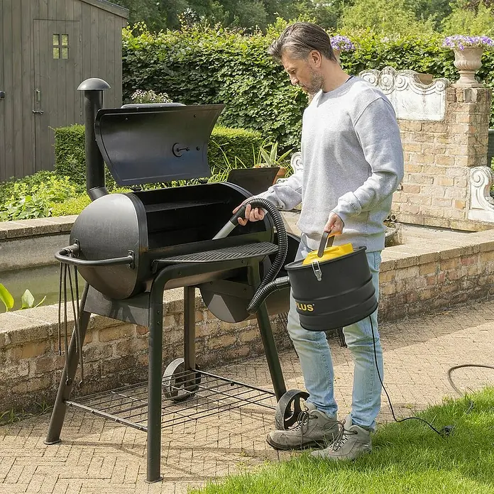 PowerPlus Aspirador de cenizas eléctrico POWX3013 - 1.600 W, Volumen de recipiente: 20 l, Sin bolsa - Un hombre limpiando una parrilla con un aspirador de cenizas negro con detalles amarillos en el jardín.