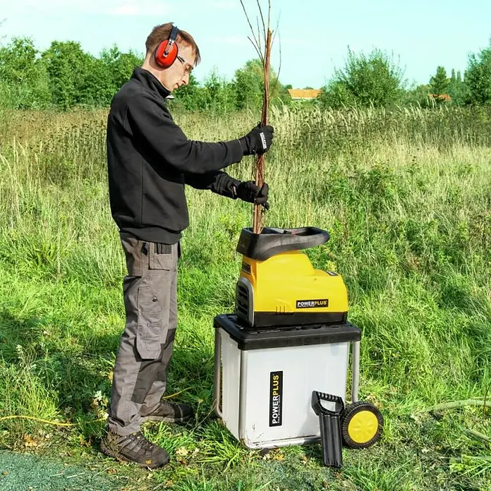 PowerPlus Biotrituradora eléctrica de rodillo POWXG6462 - Grosor de rama máx.: 4,5 cm, 2.800 W - Un hombre está usando una trituradora de jardín POWERPLUS al aire libre para triturar ramas. El dispositivo se coloca en un contenedor para recoger el material triturado.