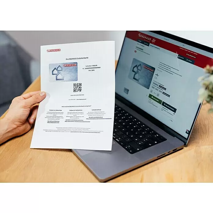 Gutscheinkarte - Zum Selbstausdruck, 50 Euro, Konglomerat - Eine Hand hält einen Ausdruck einer Bauhaus Gutscheinkarte mit Guthaben, Kartennummer und PIN neben einem Laptop mit der Bauhaus Webseite.