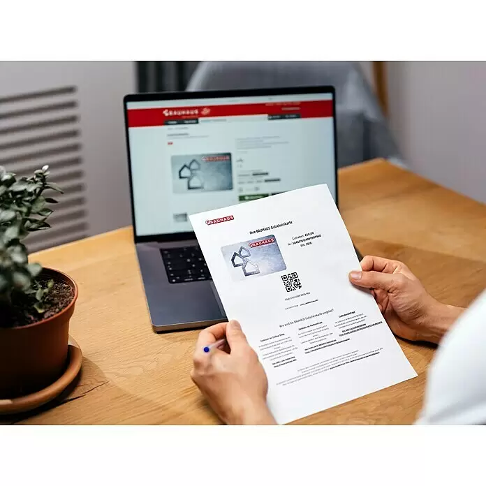 Gutscheinkarte - Zum Selbstausdruck, 50 Euro, Konglomerat - Eine Person hält ein gedrucktes BAUHAUS Gutscheinkartendokument mit Wert und Einlösedetails, im Hintergrund ein Laptop mit der BAUHAUS Website.