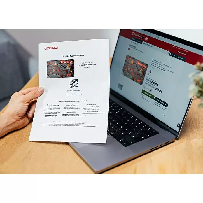 Gutscheinkarte - Zum Selbstausdruck, Individueller Betrag, Grillkohle - Person hält Bauhaus Geschenkgutschein (€60) mit Holzkohle-Motiv, daneben Laptop mit der Produktseite des Gutscheins.