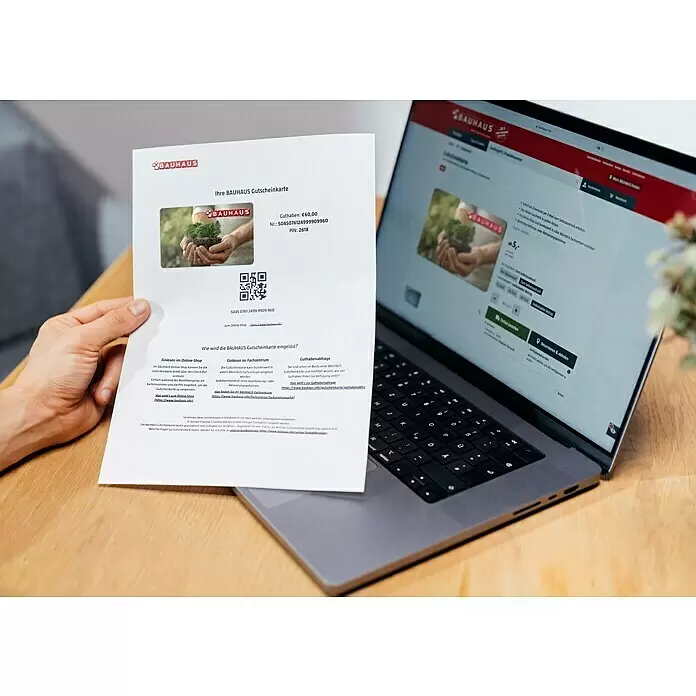 Gutscheinkarte - Zum Selbstausdruck, Individueller Betrag, Bäume - Eine Person hält einen Ausdruck einer Bauhaus Geschenkkarte neben einem Laptop, der dieselbe Geschenkkarte im Bauhaus Online-Shop anzeigt.