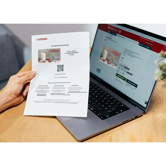 Gutscheinkarte - Zum Selbstausdruck, 50 Euro, Weihnachtsstern - Eine Hand hält einen Bauhaus Gutschein, der auf einem Laptop vor der Bauhaus-Website liegt. Der Gutschein hat ein Guthaben von 60 €.