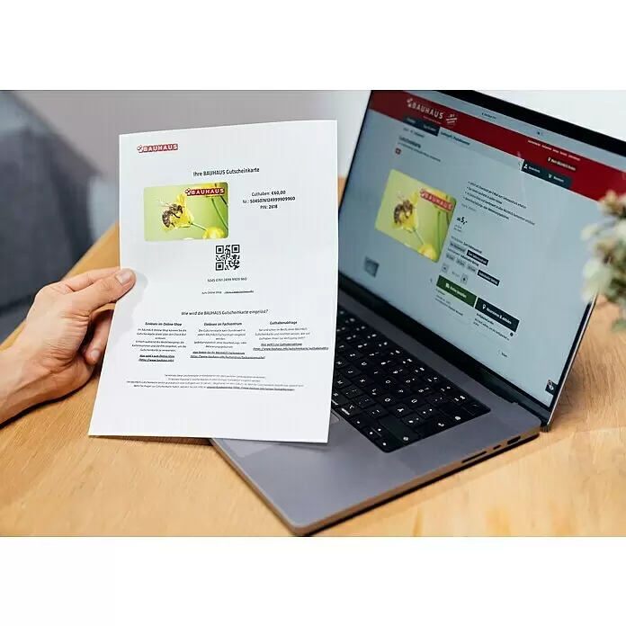 Gutscheinkarte - Zum Selbstausdruck, 10 Euro, Biene - Eine Person hält einen ausgedruckten Bauhaus-Geschenkgutschein mit einer Biene, neben einem Laptop, der die digitale Version auf der Website anzeigt.