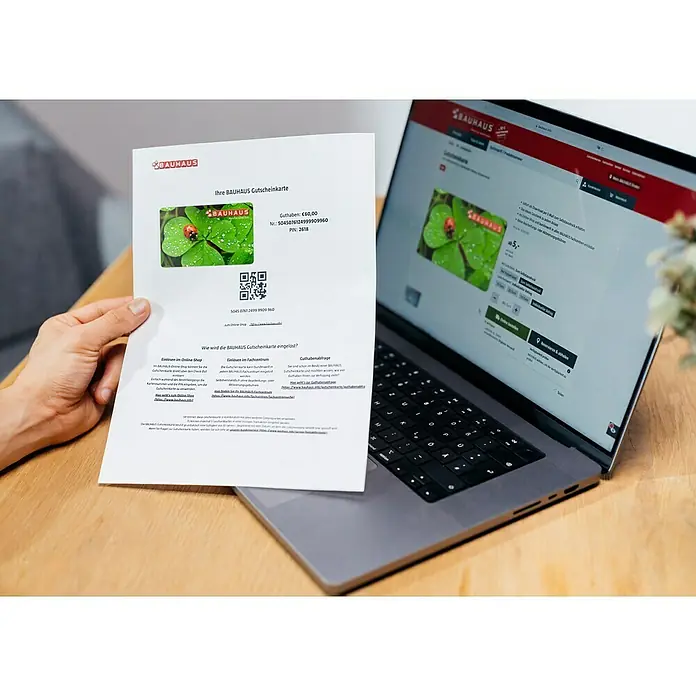 Gutscheinkarte - Zum Selbstausdruck, 10 Euro, Kleeblatt - Einlösen eines Bauhaus-Gutscheins online: Der Gutschein im Wert von 60,00€ mit einem QR-Code und einer PIN-Nummer wird neben einem Laptop gehalten.