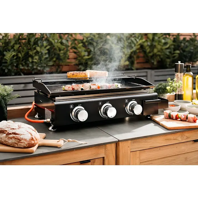 Kingstone Barbacoa de gas Nebraska - Superficie de la parrilla (An x Pr): 63,5 x 31 cm, Número de quemadores: 3 - Una parrilla al aire libre con comida encima, rodeada de condimentos y utensilios. El foco está en la parrilla y la preparación.