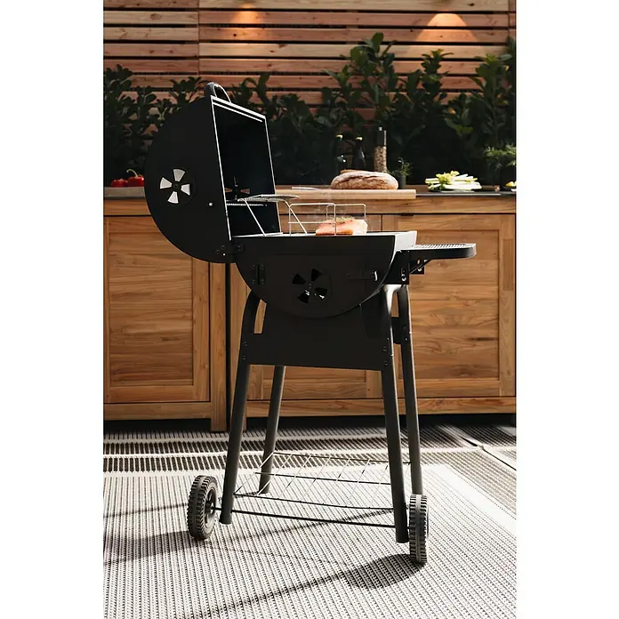 Grillstar Holzkohlegrill Grilltonne - B x T x H: 68 x 66 x 109 cm, Grillfläche (B x T): 46 x 43 cm, Schwarz - Ein schwarzer Holzkohlegrill mit offenem Deckel und einem Stück Fisch darauf, steht auf einem gemusterten Teppich vor einem Holzzaun.
