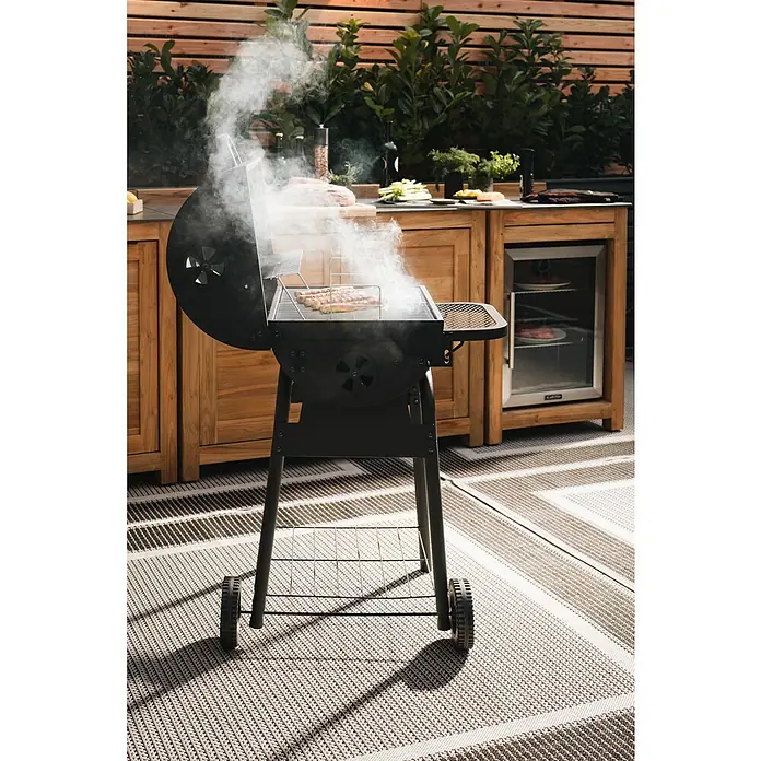 Grillstar Holzkohlegrill Grilltonne - B x T x H: 68 x 66 x 109 cm, Grillfläche (B x T): 46 x 43 cm, Schwarz - Grill mit Rauchentwicklung auf einer Terrasse. Der Holzkohlegrill steht auf einem gemusterten Teppich vor einem Gartenmöbelset aus Holz.