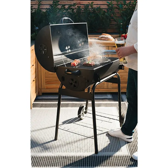 Grillstar Holzkohlegrill Grilltonne - B x T x H: 109 x 71 x 87 cm, Grillfläche (B x T): 64 x 43 cm, Schwarz - Ein Holzkohlegrill steht auf einer Terrasse, während jemand Fleisch und Gemüse grillt und ein Bier genießt.