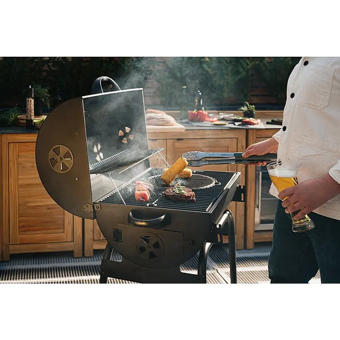 Kingstone Grillzange - Länge: 46 cm - Bild von einem Holzkohlegrill, auf dem Essen gegrillt wird, mit einer Person, die ein Bier hält. Der Fokus liegt auf dem Outdoor-Grillerlebnis.