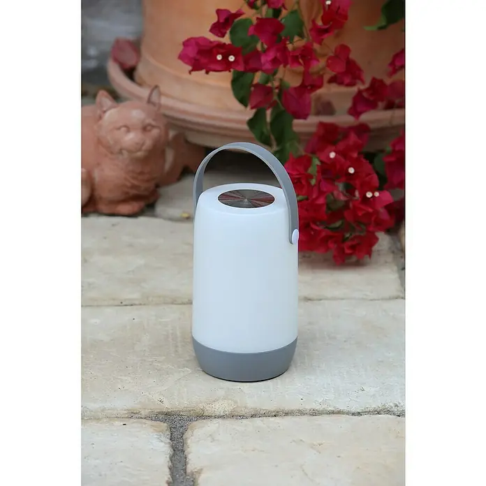 Mobiles Licht Sona Touch - Weiß, Höhe: 19 cm, IP44 - Nahaufnahme einer tragbaren LED-Laterne mit einem grauen Griff und einer grauen Basis, die auf einem gepflasterten Gehweg mit Blumen im Hintergrund steht.