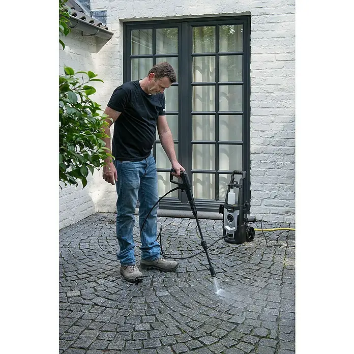 PowerPlus Hidrolimpiadora de alta presión POWXG90405 - 110 bar, 1.400 W, Capacidad de bombeo: 390 l/h - Un hombre usa una hidrolimpiadora para limpiar un área pavimentada exterior, con la hidrolimpiadora colocada cerca.