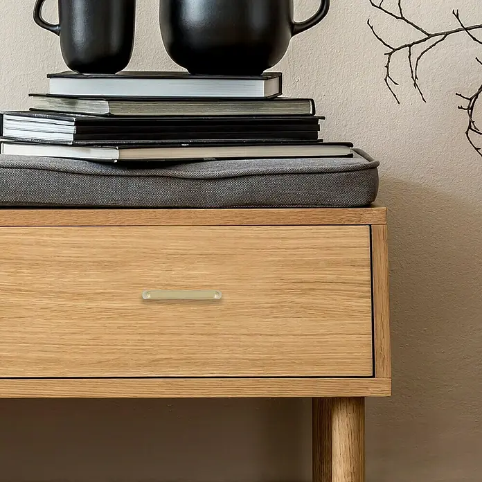 Häfele Tirador para muebles - Tipo de tirador del mueble: Estribo, Acero, Distancia entre orificios: 64 mm - Primer plano de una cómoda de madera con un tirador alargado y transparente sobre un fondo liso.