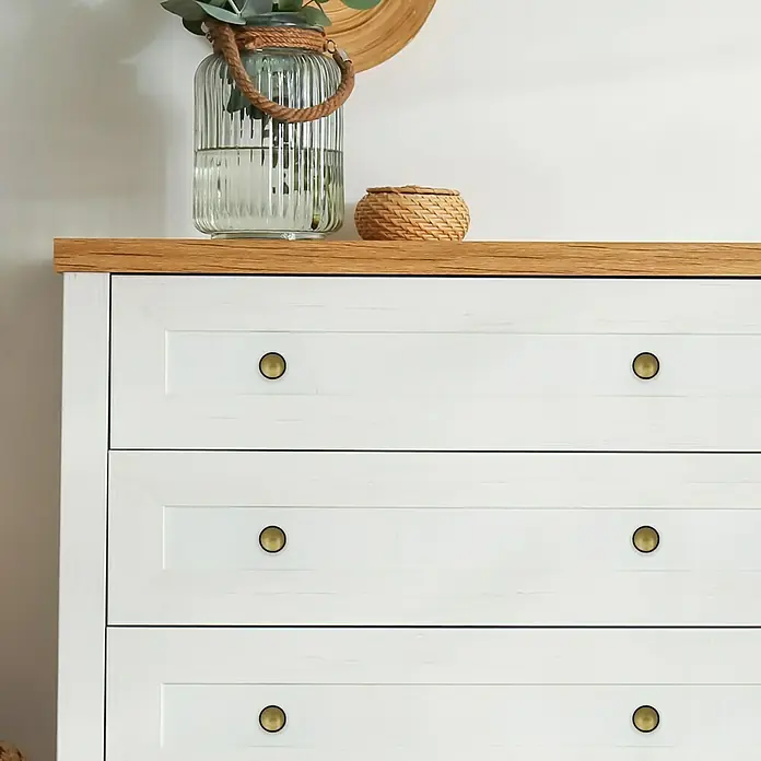 Häfele Pomo para muebles - Tipo de tirador del mueble: Botón, Ø x L: 30 x 25 mm - Primer plano de una cómoda blanca con tapa de madera y pomos dorados, con un jarrón de cristal y una cesta pequeña encima.