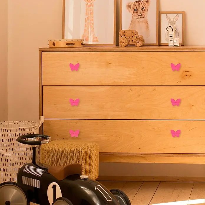 Häfele Pomo infantil para mueble - Botón, Ø x Al: 70 x 53 mm, Lila, Mariposa - En la imagen se ve una cómoda de madera adornada con pomos de mariposa rosas, que añaden un toque caprichoso y apto para niños a los muebles.