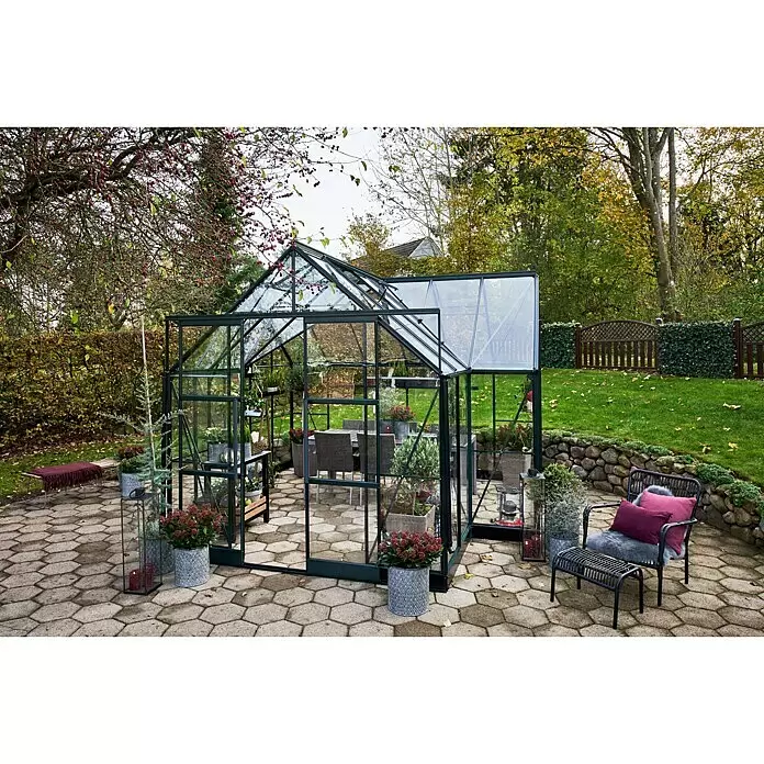 Halls Gewächshaus Garden Room - Außenmaß inkl. Dachüberstand (B x T): 389 x 390 cm, Farbe Gestell: Grün, Wandstärke: 3 mm - Ein geräumiges, dunkel gerahmtes Gewächshaus mit transparenten Paneelen auf einer Terrasse, umgeben von Gartenmöbeln und Topfpflanzen.