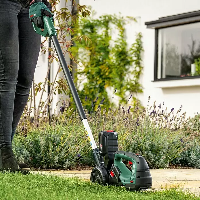 Bosch Power for All 18V Teleskopstiel - Passend für: AdvancedShear 18V-10 - Eine Person trimmt mit einem grünen und schwarzen Bosch Akku-Rasentrimmer mit Rädern Gras entlang eines Gartenwegs.