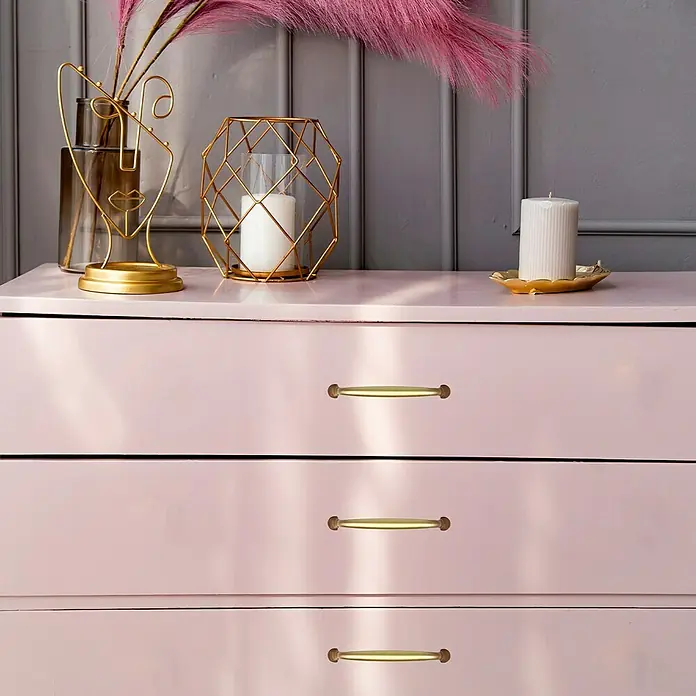 Häfele Tirador para muebles - Tipo de tirador del mueble: Estribo, Distancia entre orificios: 128 mm - Primer plano de una cómoda rosa con tres cajones y tiradores dorados, decorada con velas y elementos decorativos.