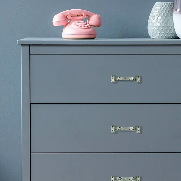 Häfele Tirador para muebles - An x L: 50 x 100 mm - Primer plano de una cómoda azul grisácea con tres cajones y tiradores de plata ornamentados, coronada con un teléfono rosa y un jarrón gris.
