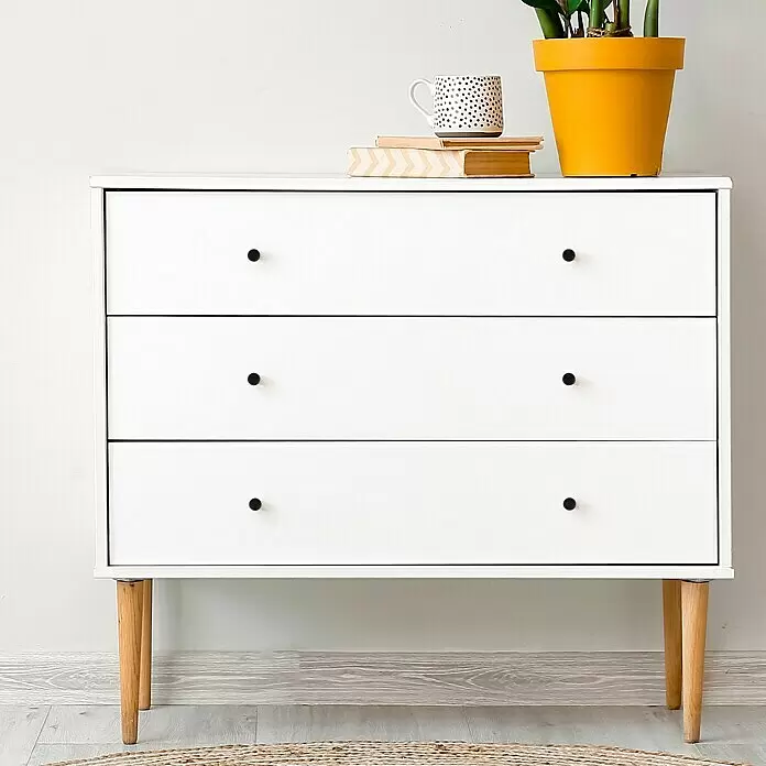 Häfele Pomo para muebles - Tipo de tirador del mueble: Botón - Cómoda blanca de tres cajones con pomos negros y patas de madera clara, con una planta, taza y libros encima.