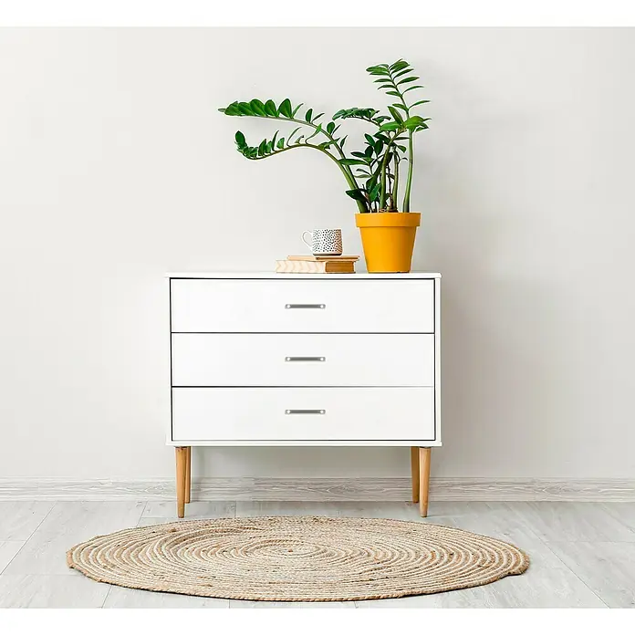 Häfele Möbelgriff - Typ Möbelgriff: Bügel, L x B x H: 102 x 12 x 30 mm, Lochabstand: 96 mm - Helle Kommode mit drei Schubladen, darauf eine Zamioculcas-Pflanze in einem gelben Topf, Bücher und eine Tasse. Darunter liegt ein runder Teppich.
