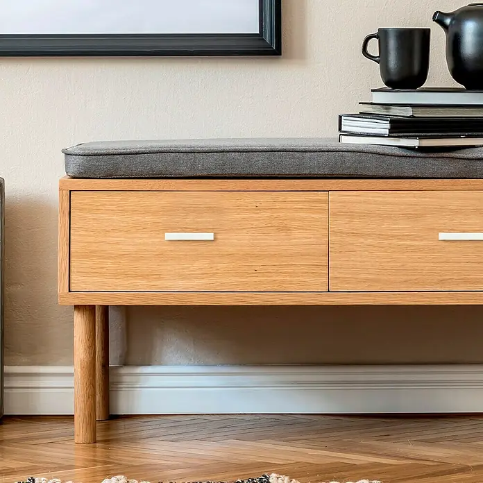 Möbelgriff - Typ Möbelgriff: Bügel, L x B x H: 138 x 21 x 26 mm, Aluminium - Diese Aufnahme zeigt eine Bank aus hellem Holz mit zwei Schubladen und einer grauen Sitzfläche. Bücher und ein Teeservice stehen darauf.