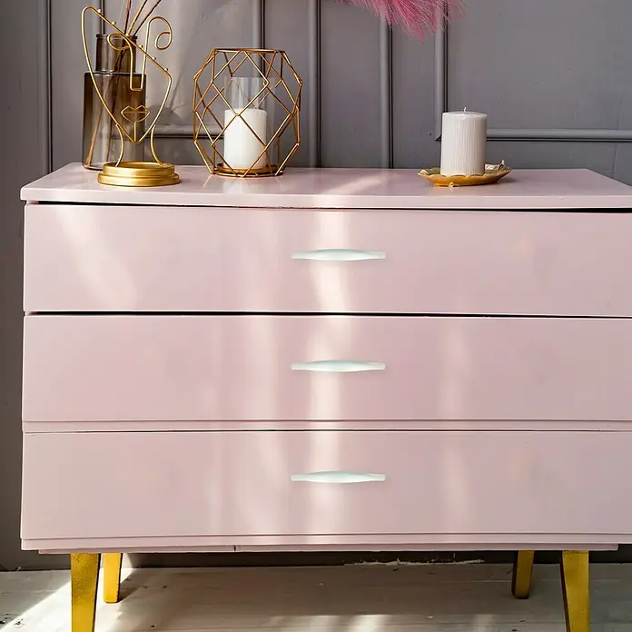 Möbelgriff - Typ Möbelgriff: Bügel, L x B x H: 154 x 19 x 30 mm, Sonstige, Verchromt - Nahaufnahme einer rosa Kommode mit drei Schubladen und weißen Griffen, die goldene Beine hat, und dekorative Gegenstände auf der Oberseite.