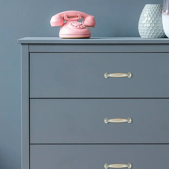Möbelgriff - Typ Möbelgriff: Bügel, Silber - Nahaufnahme einer blaugrauen Kommode mit drei Schubladen und verzierten Griffen. Auf der Kommode befinden sich ein rosa Telefon und eine dekorative Vase.