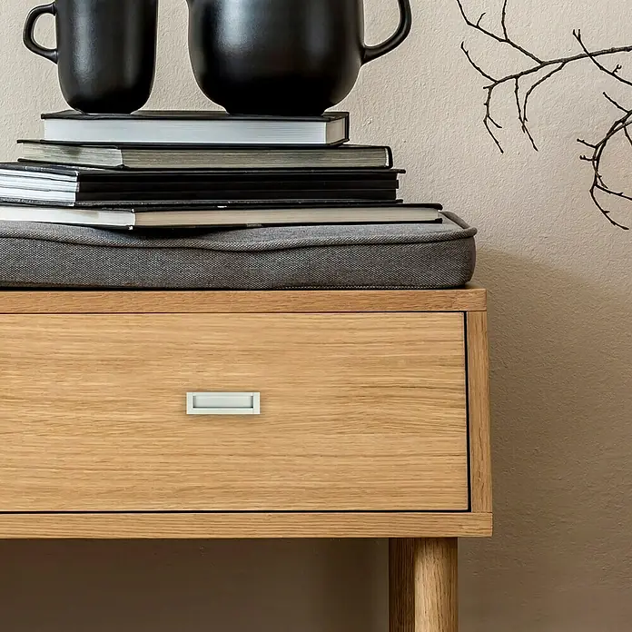 Häfele Tirador para muebles - Tipo de tirador del mueble: Concha, Aluminio - Primer plano de una mesita de noche marrón claro con un cajón con un elegante tirador plateado. Decorada con tazas oscuras y libros.