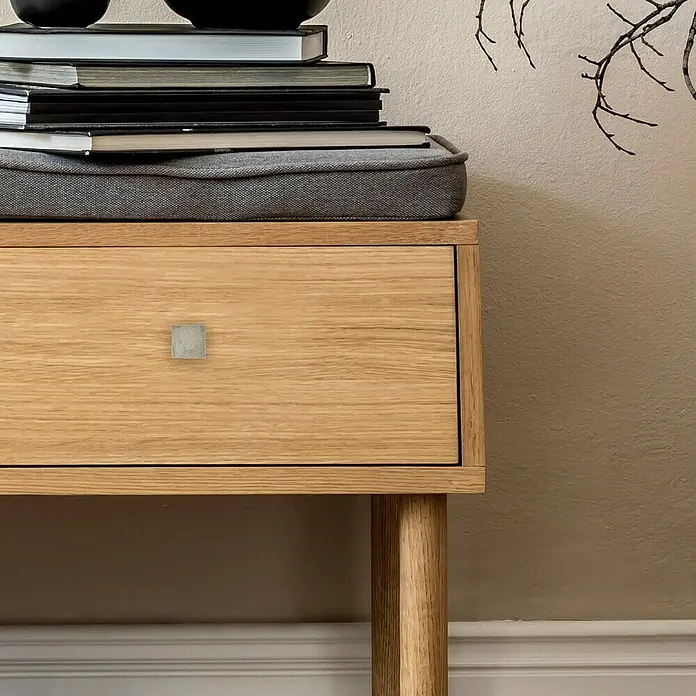 Häfele Pomo para muebles - Tipo de tirador del mueble: Botón, Niquelado, Gris - Primer plano de una mesita de noche de roble con un cajón con tirador cuadrado, sobre la que se apoyan libros y adornos.