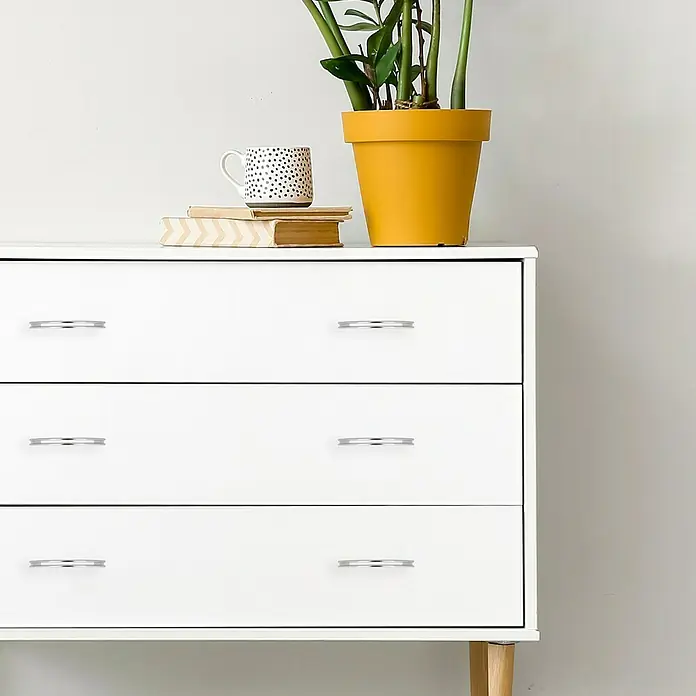 Häfele Tirador para muebles - Tipo de tirador del mueble: Estribo, L x An x Al: 124 x 12 x 28 mm, Otros, Cromado, Distancia entre orificios: 96 mm - Aparador blanco con tres cajones y tiradores plateados, decorado con plantas, una taza y libros para un ambiente de estar elegante.