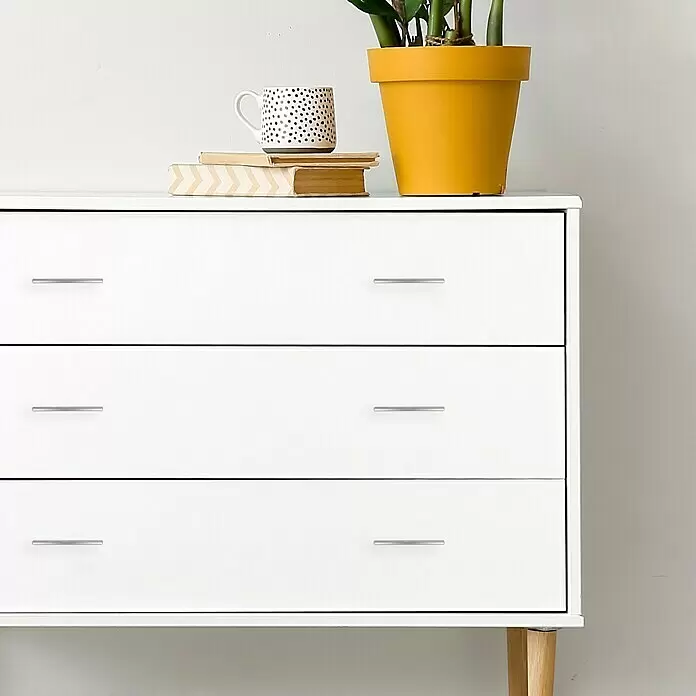 Häfele Tirador para muebles - Niquelado, Distancia entre orificios: 64 mm, An x L: 30 x 70 mm - Cómoda blanca de tres cajones con tiradores plateados y patas de madera clara, decorada con libros, taza y planta encima.