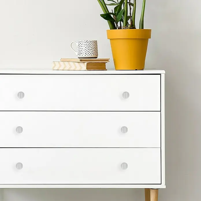 Häfele Pomo para muebles - Tipo de tirador del mueble: Botón, Ø x Al: 25 x 27 mm, Brillante - Primer plano de una cómoda blanca con tres cajones y pomos redondos transparentes, decorada con plantas, libros y una taza.